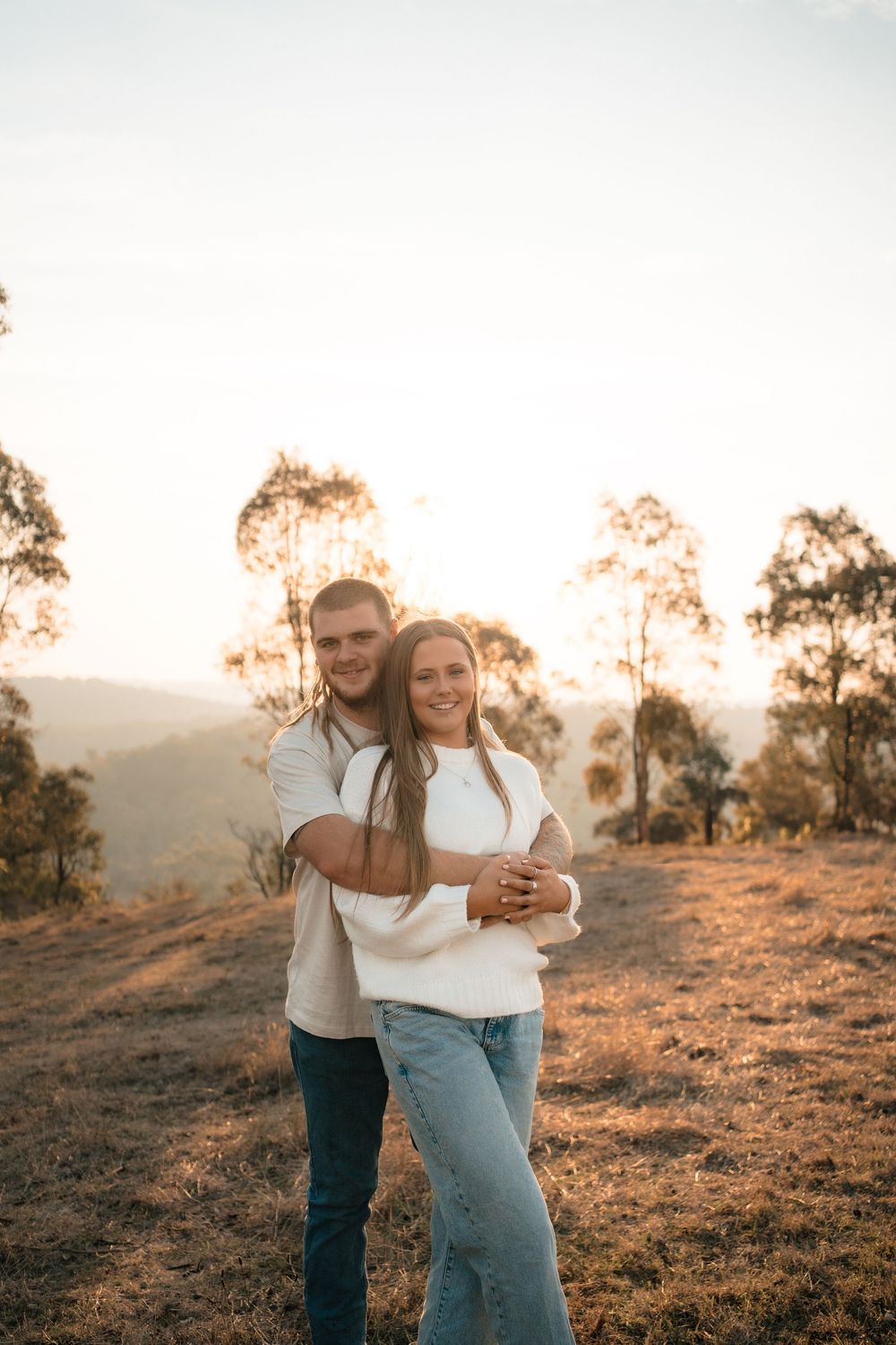 Couple embracing during golden hour engagement photoshoot in scenic countryside setting.