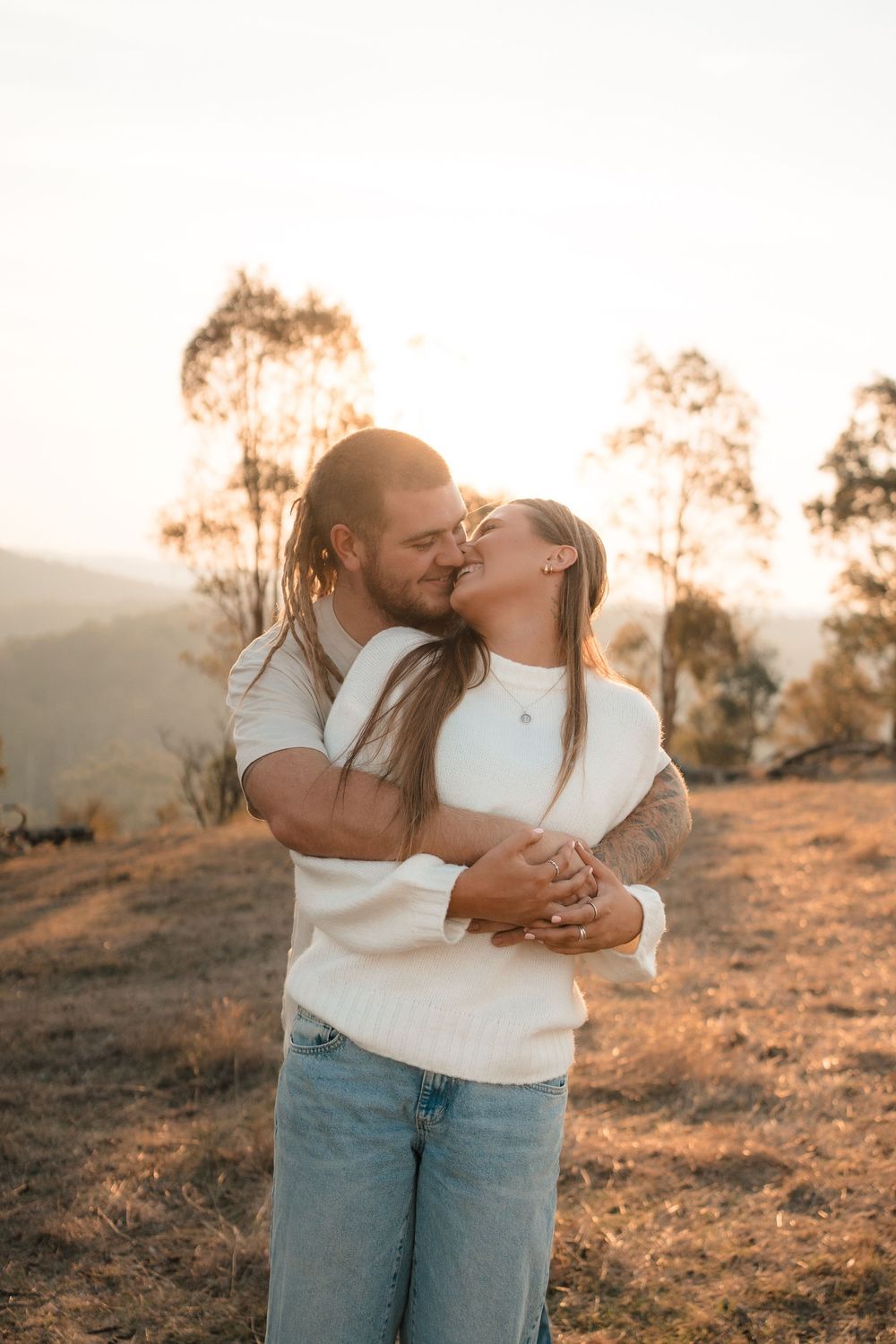 A couple embraces and kisses during golden hour on a hilltop with eucalyptus trees in the background.