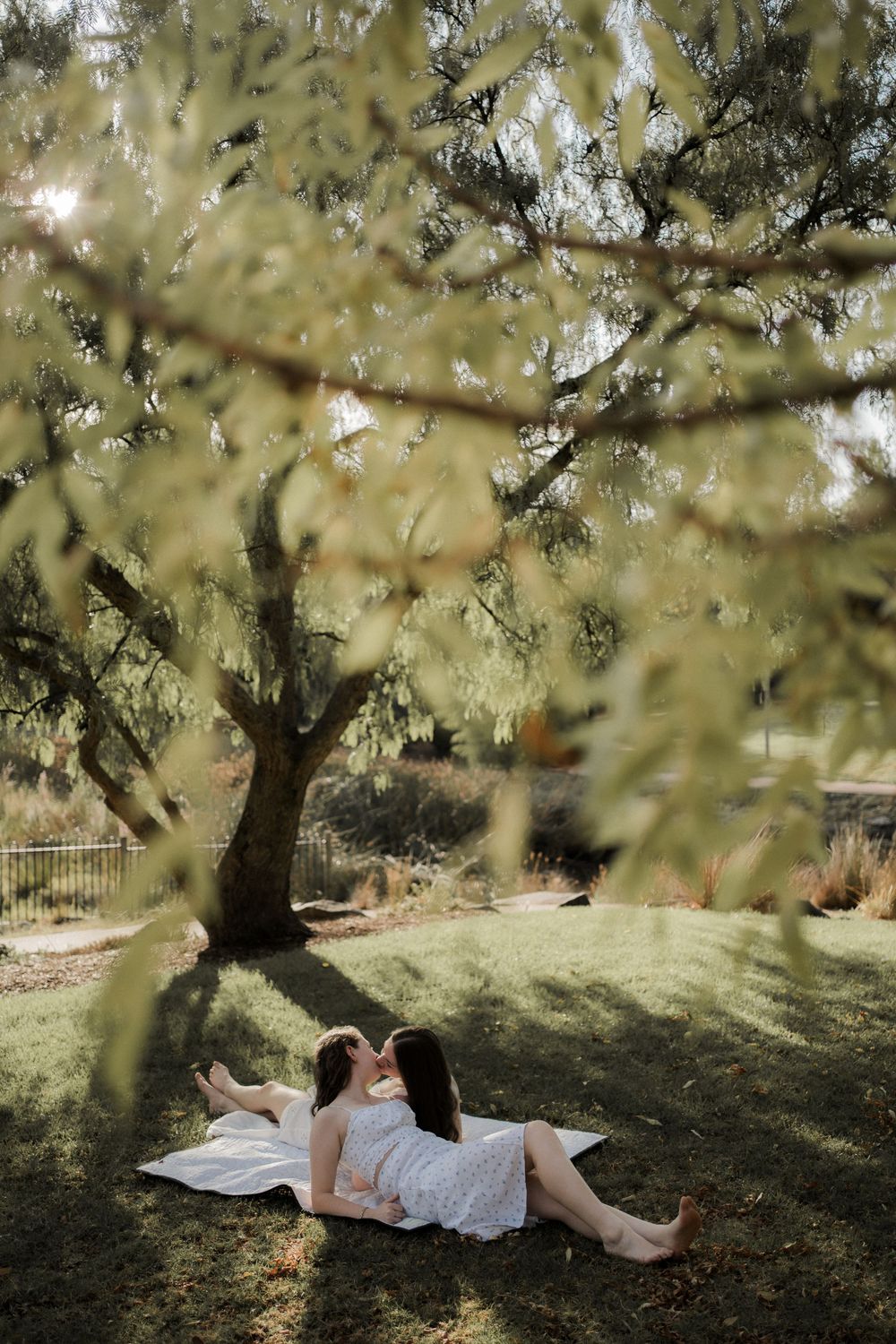 Two people relax on a blanket under the dappled shade of a large tree in a peaceful garden setting.