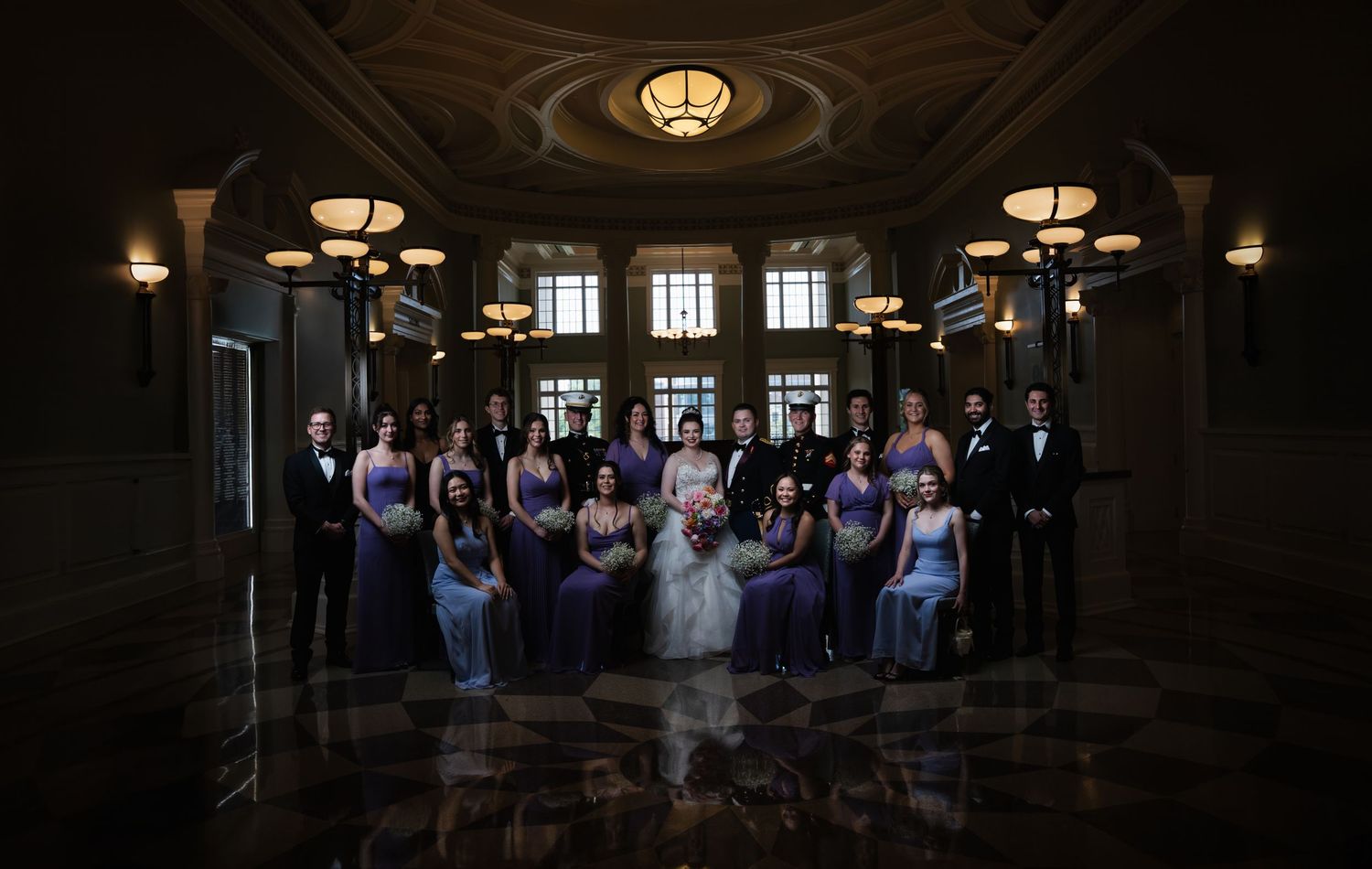 A wedding party poses dramatically in a dark elegant hallway with ornate ceiling and lighting fixtures.