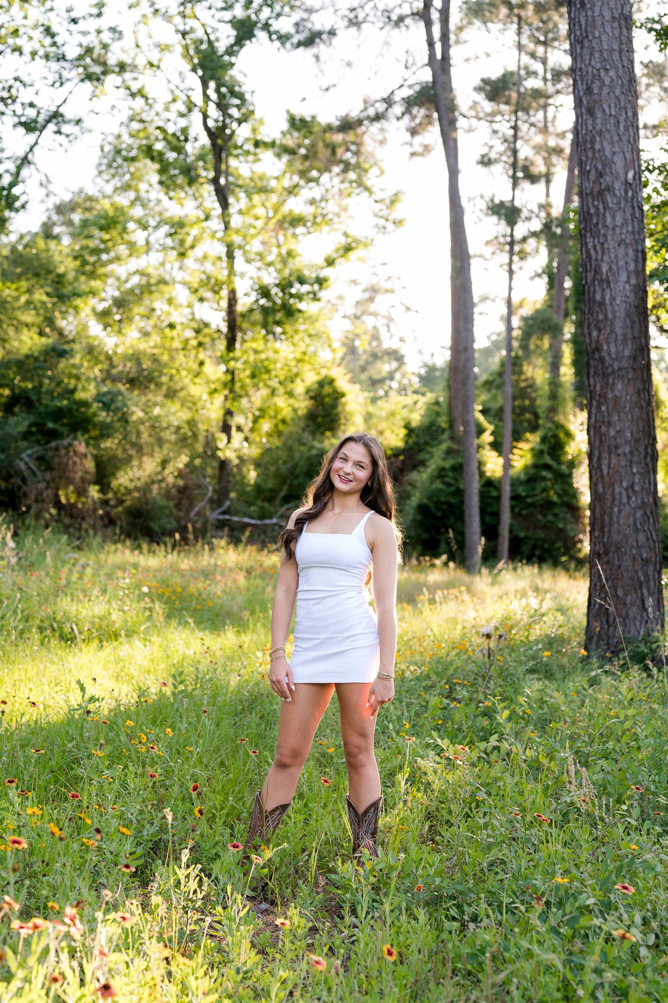 A white sundress stands out against a sunlit forest meadow with wildflowers and tall pine trees in summer.