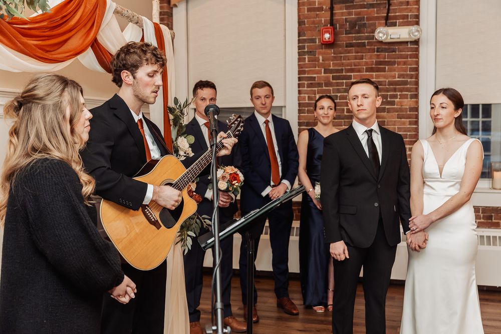 Guitarist performs at indoor wedding ceremony with exposed brick wall and orange drapery as backdrop.