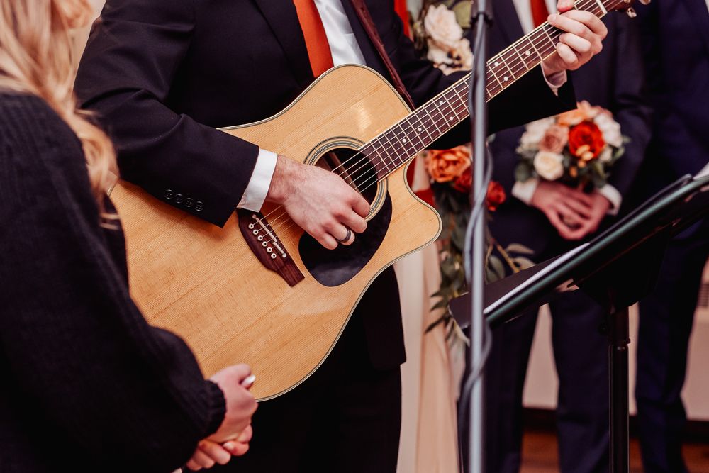 Close up of hands playing acoustic guitar during wedding ceremony with floral bouquet in background.