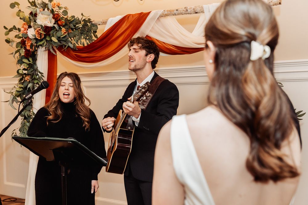 Musicians perform at a wedding ceremony under elegant draping and floral decor, with an acoustic guitar and vocalist.