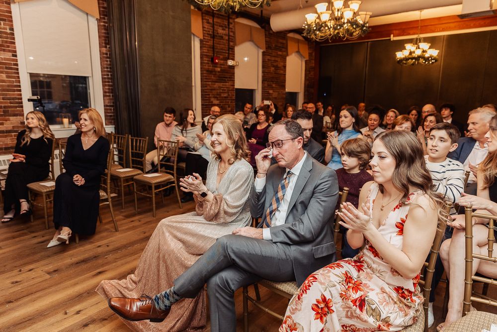 Wedding guests seated in rows of wooden chairs applaud during an indoor ceremony in a rustic venue with chandeliers.