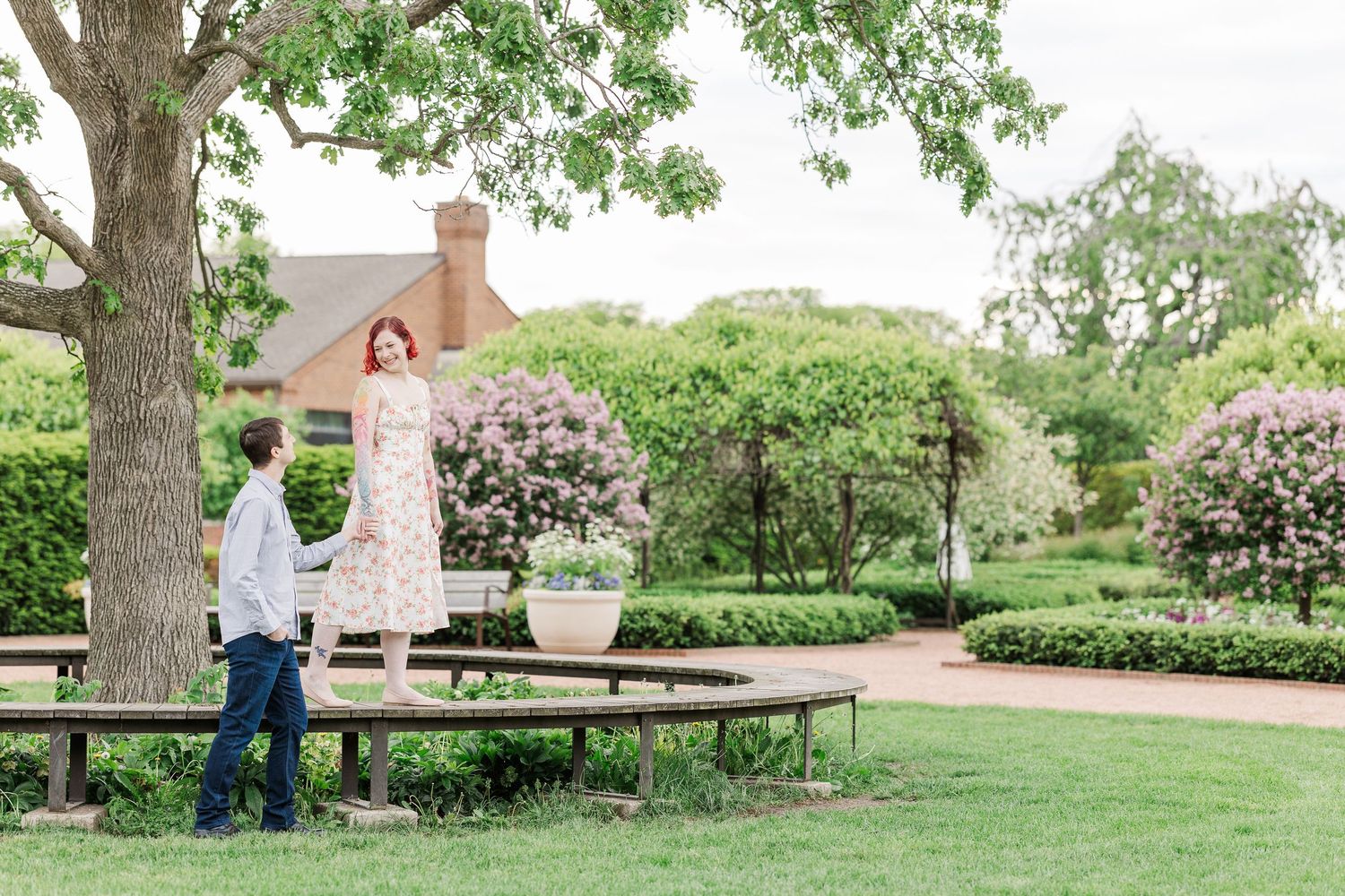 Two people share a romantic moment in a serene garden setting with blooming lilac bushes and a rustic wooden bench at Cantigny Park