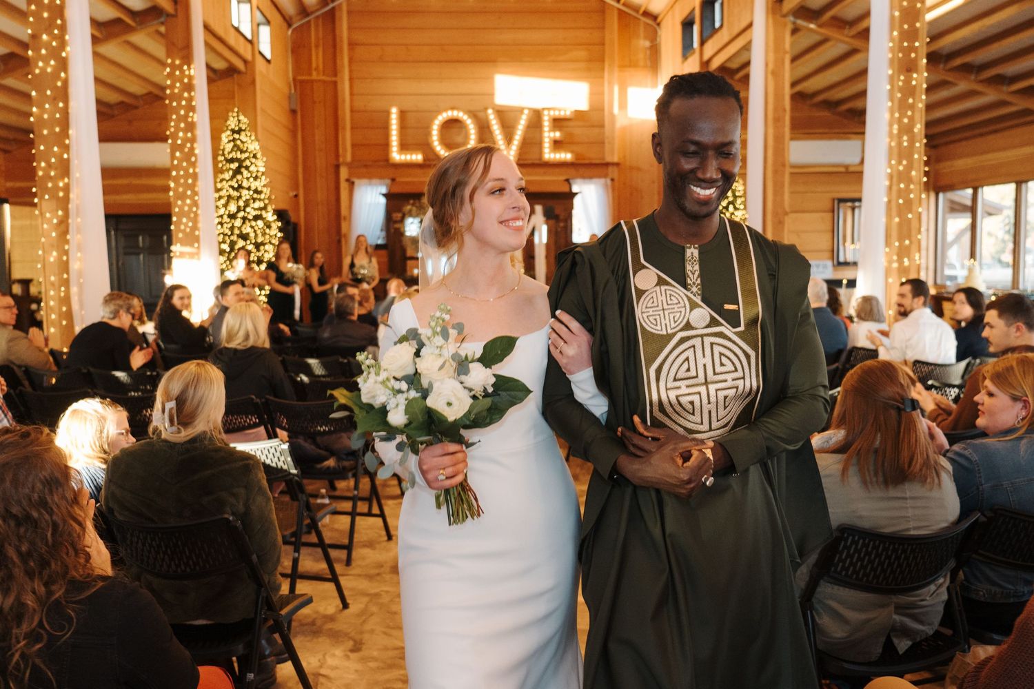 Wedding ceremony in rustic venue with illuminated LOVE sign as two people walk down aisle after exchanging vows.