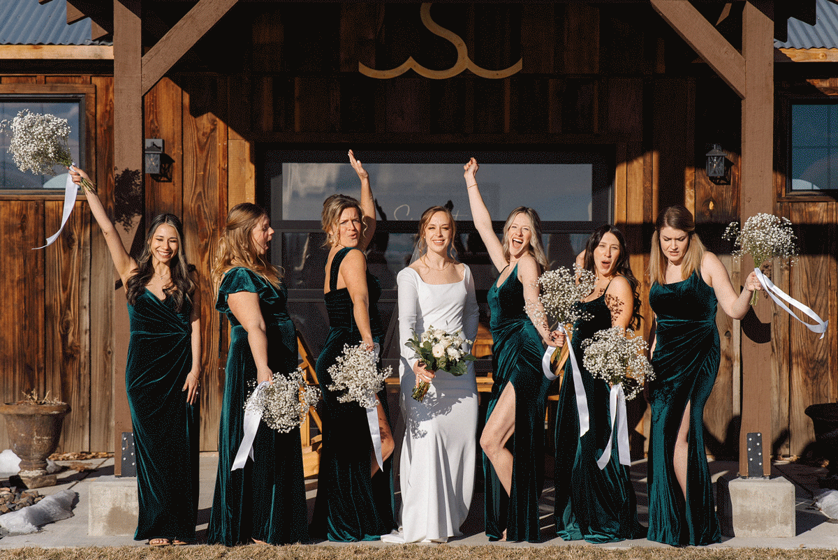 Wedding party in elegant velvet dresses poses joyfully outside rustic wooden barn in winter setting.