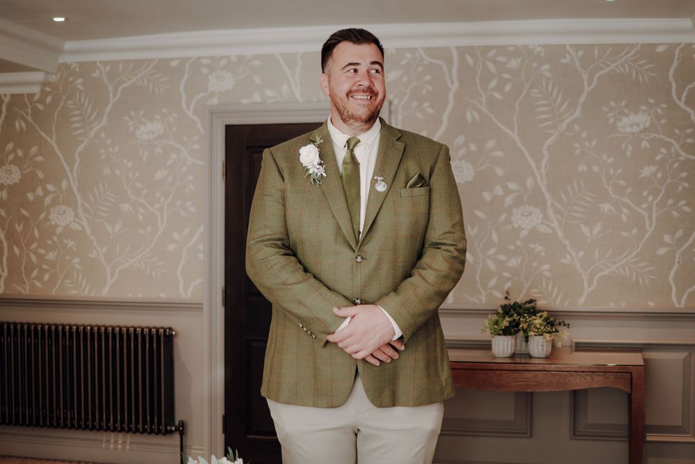 A person in an olive green suit jacket and cream pants stands in front of a patterned wallpaper at a formal event.