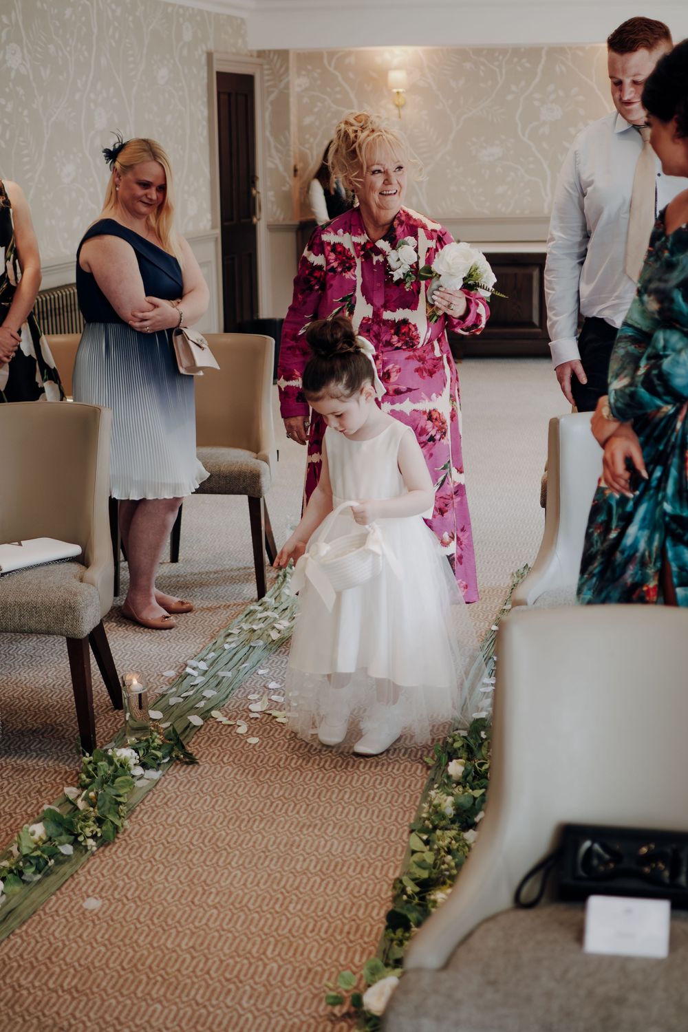 A young person in a white dress sits surrounded by decorative floral garlands on the floor during a wedding celebration.