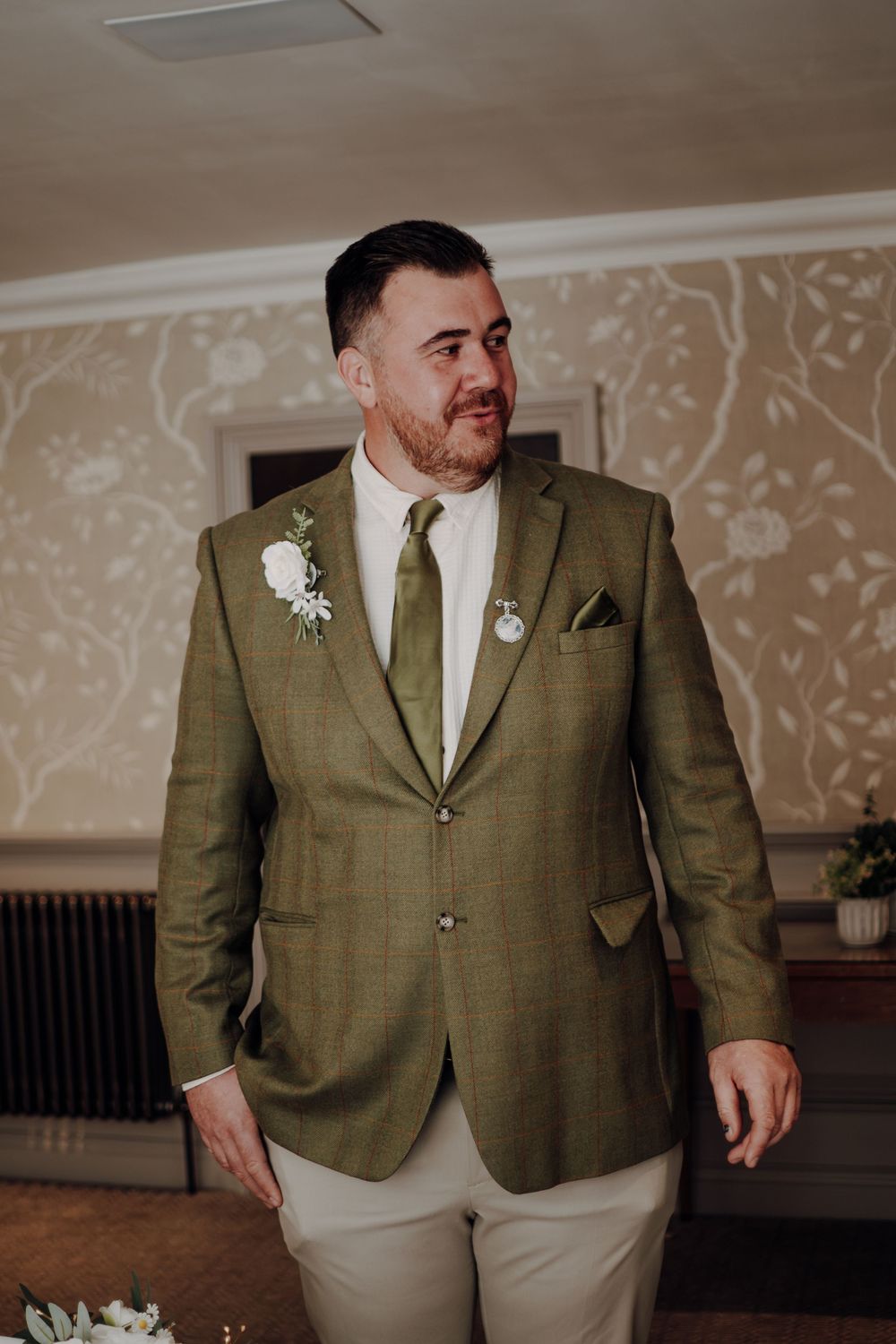 A groom wearing an olive green suit jacket with boutonniere and khaki pants at a wedding ceremony against floral wallpaper.
