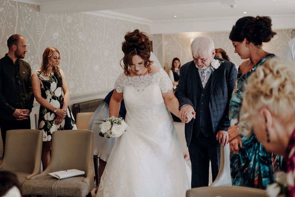 People in formal attire gather in a room as someone in a white wedding dress is escorted down the aisle.