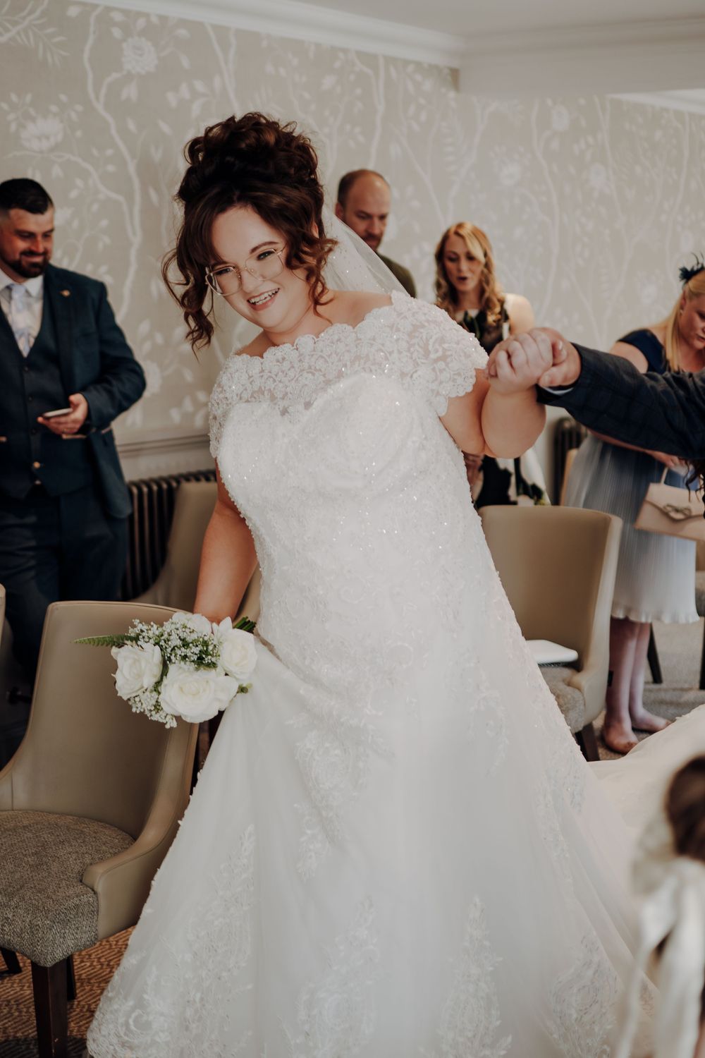 A bride in an elegant white lace wedding dress holds a small bouquet of flowers during a wedding preparation moment.
