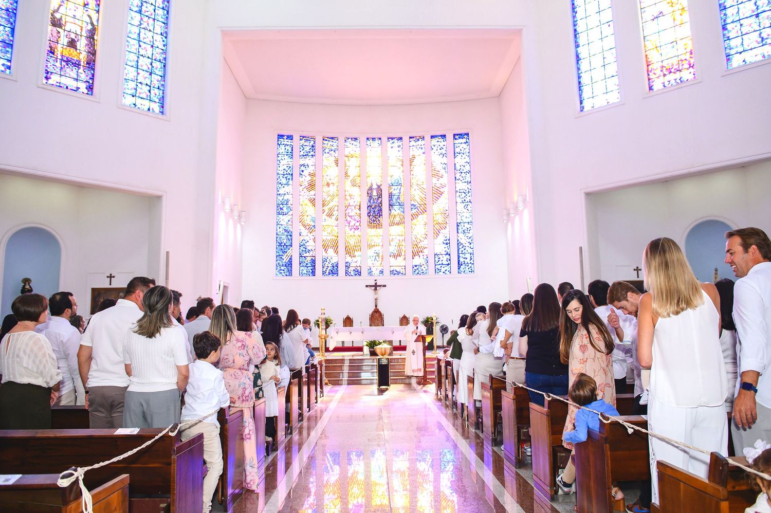 Wedding ceremony taking place in a modern church with pink walls and stained glass windows casting soft light on gathered attendees.