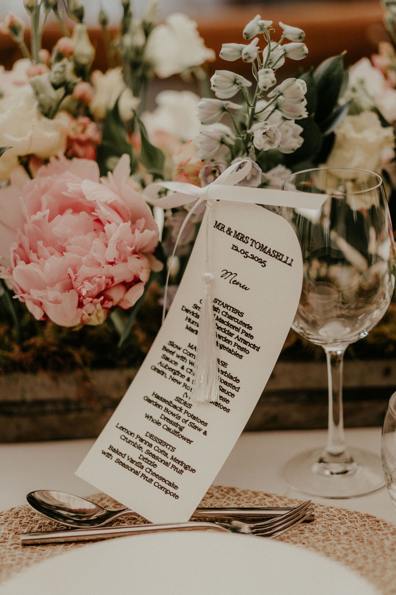 Elegant wedding menu card displayed on table with pink peony flowers, white wine glass and vintage place setting.