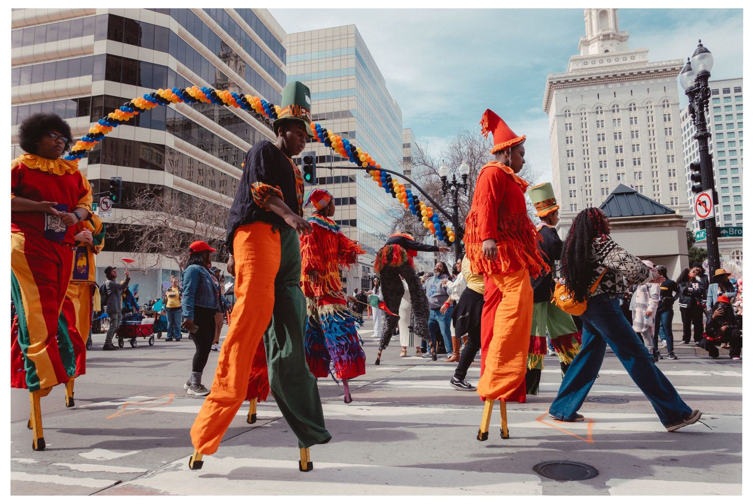 Street performers in bright orange costumes dance and entertain crowds in an urban plaza with tall buildings in the background.