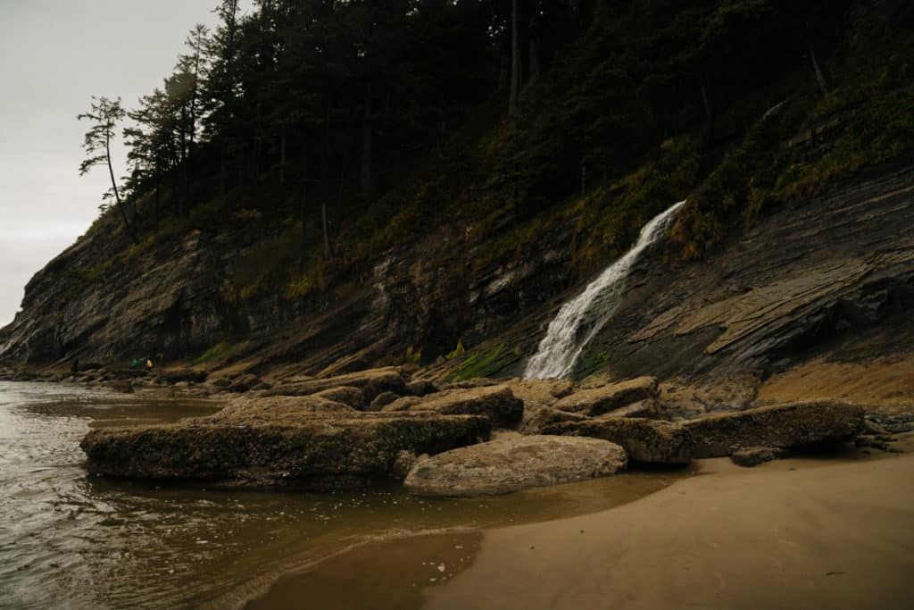 A dramatic waterfall cascades down rugged coastal cliffs onto a sandy beach with large rocks at Oregon's coastline.