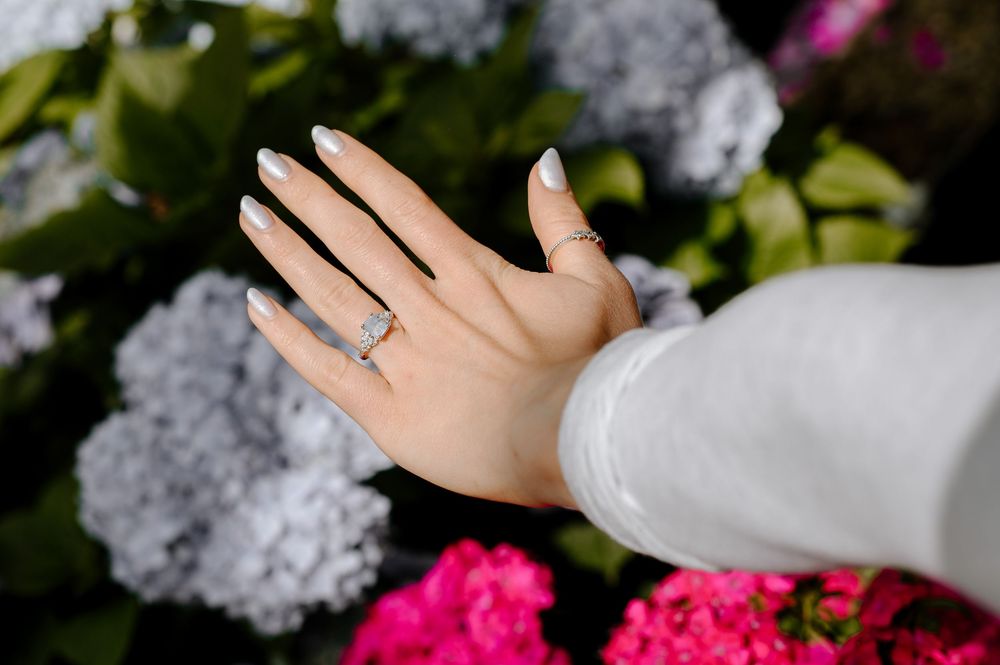 A hand wearing an engagement ring extends against a background of pink and white hydrangea flowers.