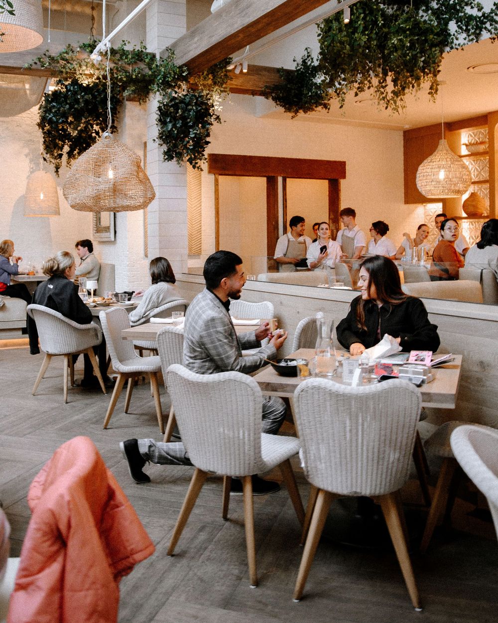 Modern restaurant interior with hanging light fixtures, lush greenery, and diners seated at white tables enjoying their meals.