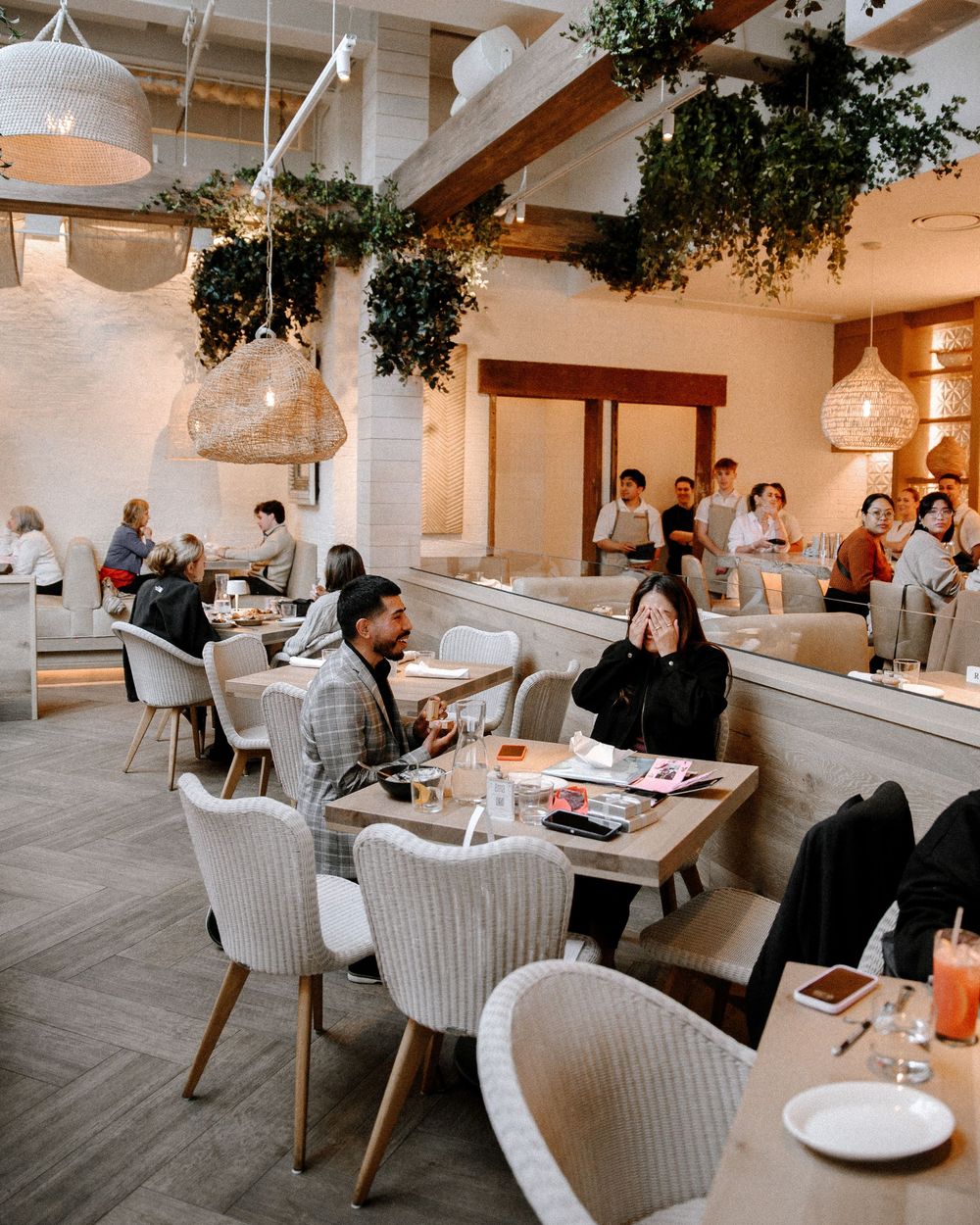 Modern restaurant interior with hanging greenery, white decor and diners enjoying meals at comfortable wicker chairs.