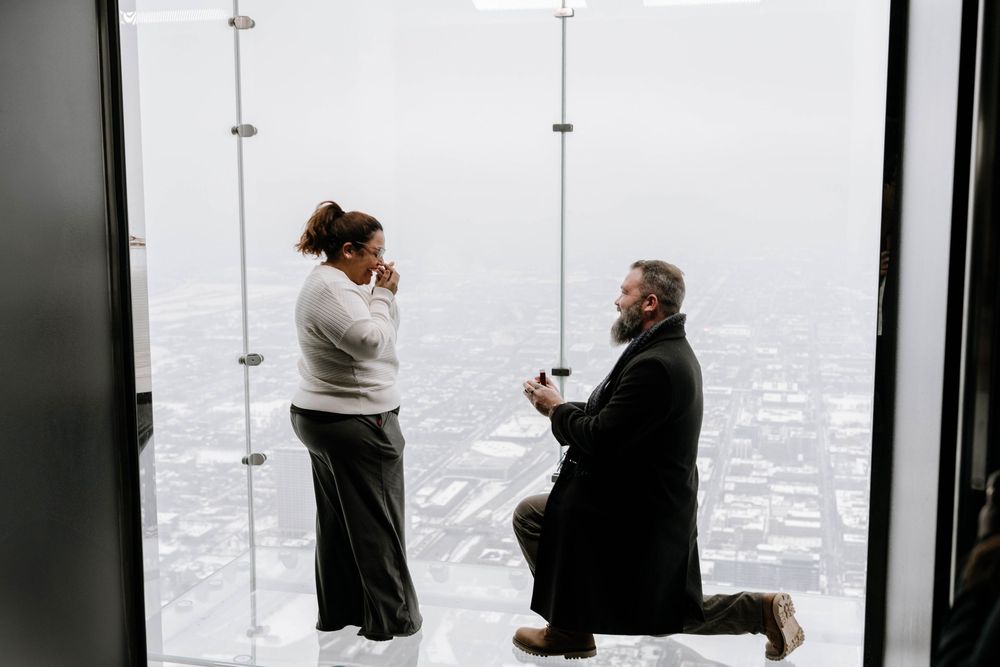 A couple shares a romantic proposal moment at an observation deck overlooking a cityscape in winter.