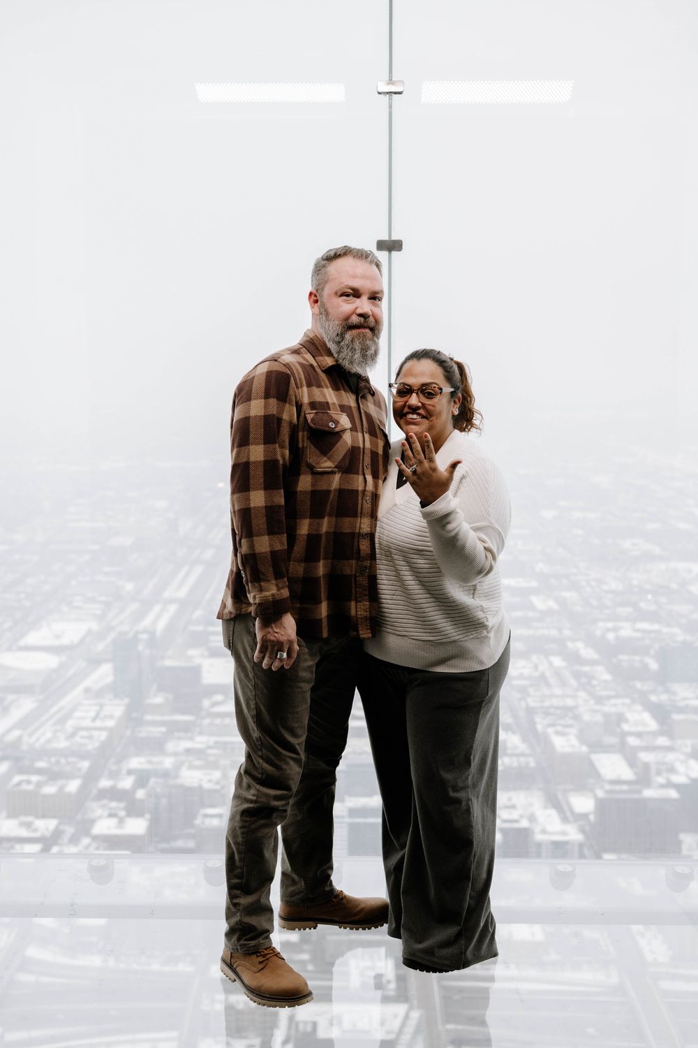 A couple stands together on a glass observation deck high above a misty cityscape below.