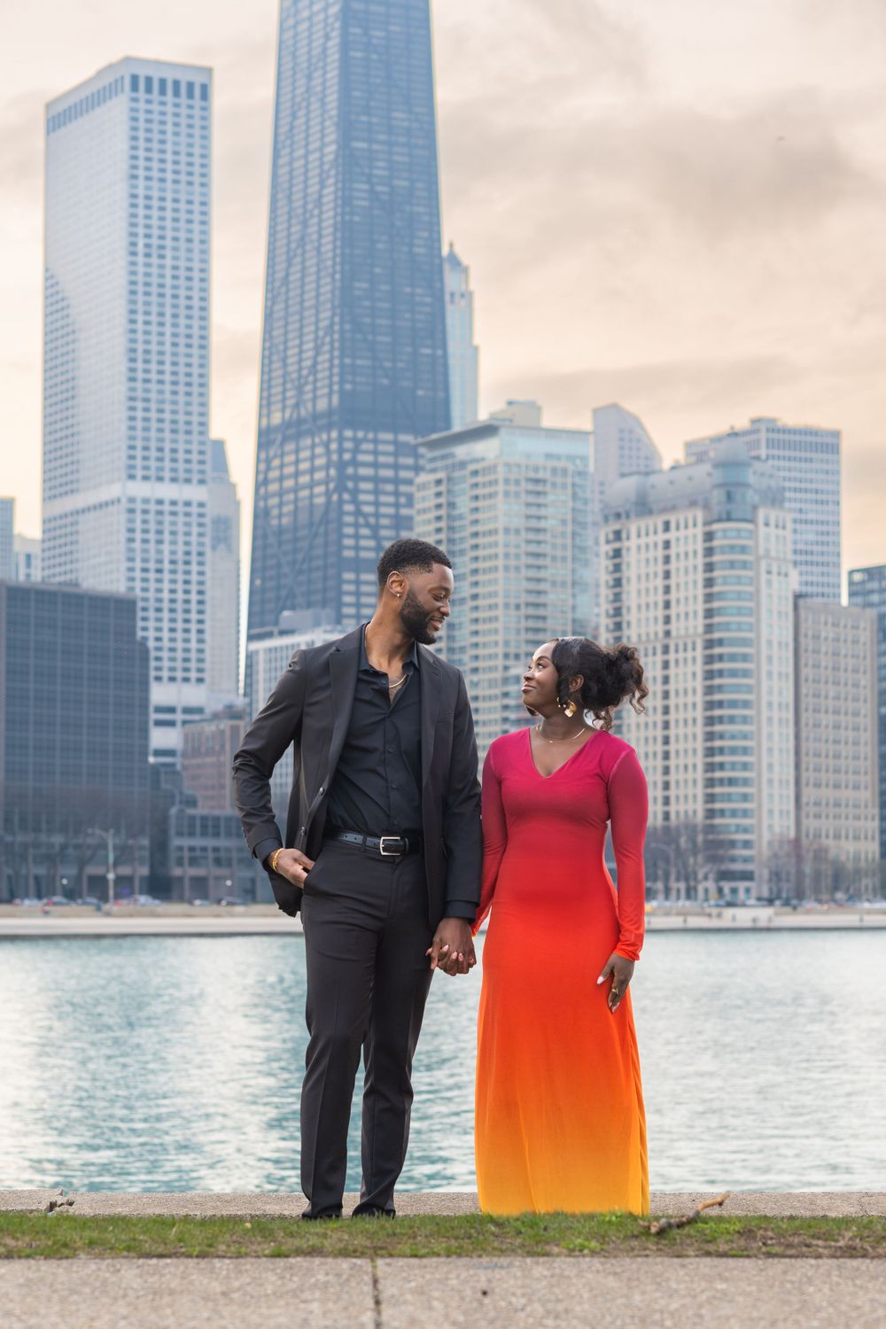 Couple in formal attire holding hands by Lake Michigan with Chicago skyline on cloudy day.