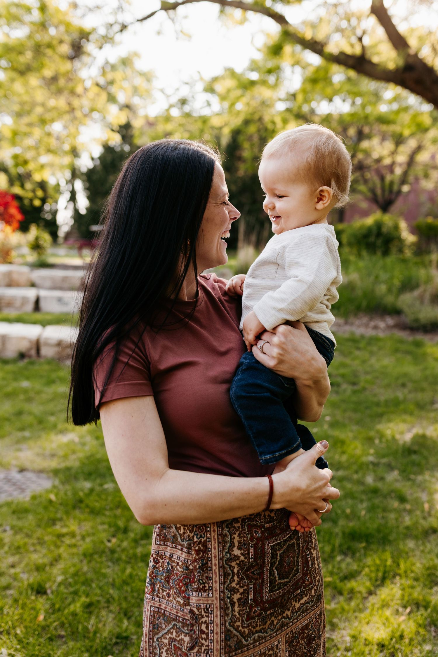 Woman in burgundy shirt holds baby in white top while sharing joyful moment together in garden setting.