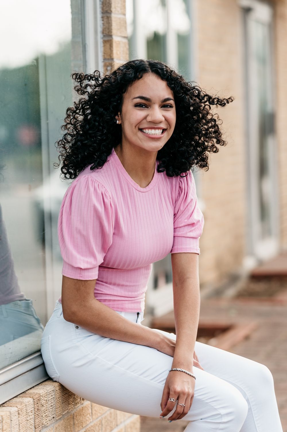 Person with natural curly hair wearing a pink ribbed top and white pants sits casually on a windowsill outdoors.