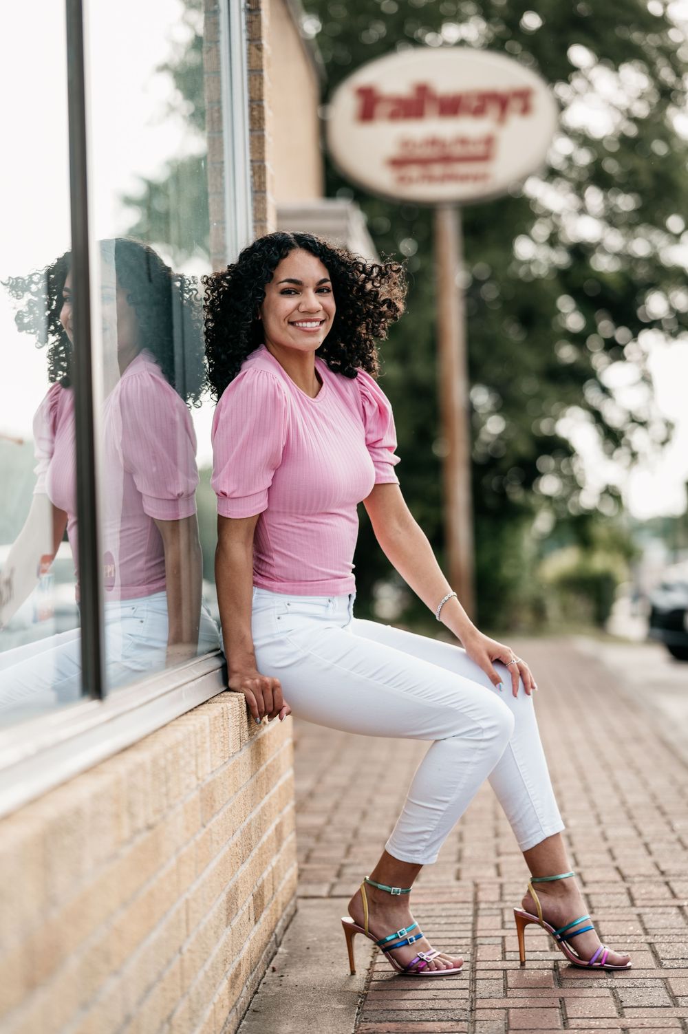 A person in a pink top and white pants sits casually on a storefront ledge with a blurred restaurant sign in the background.