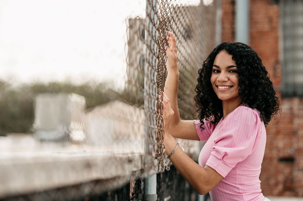 Person in a pink shirt smiles while leaning against a wire fence outdoors.