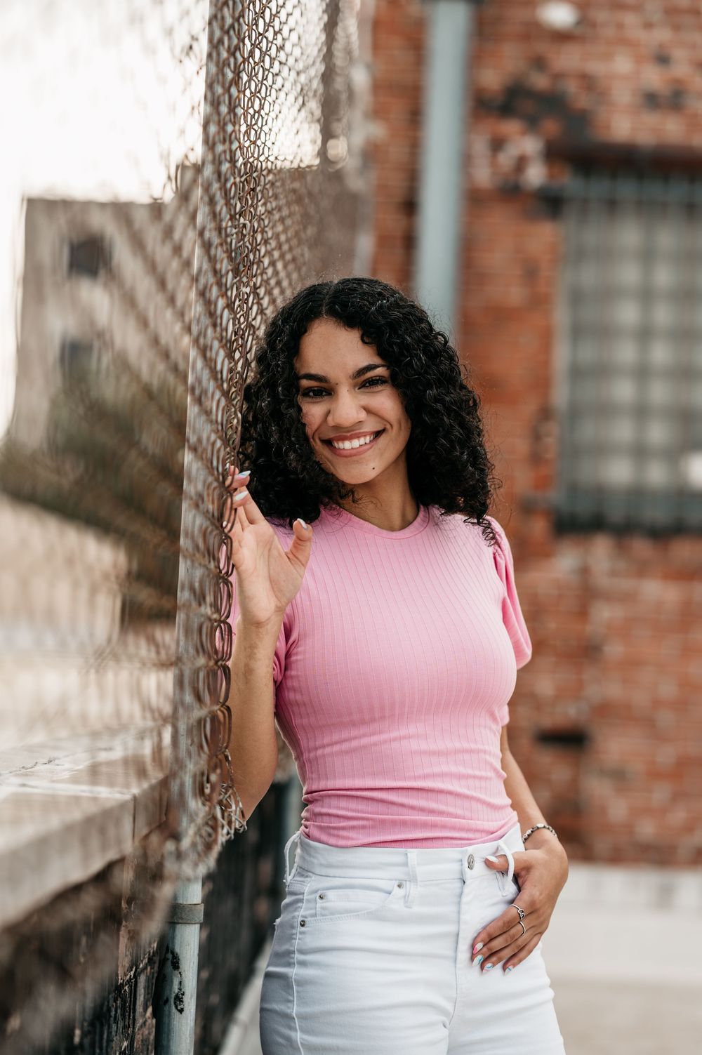 Someone in a pink ribbed top and white jeans poses casually against a chain link fence near a brick wall.