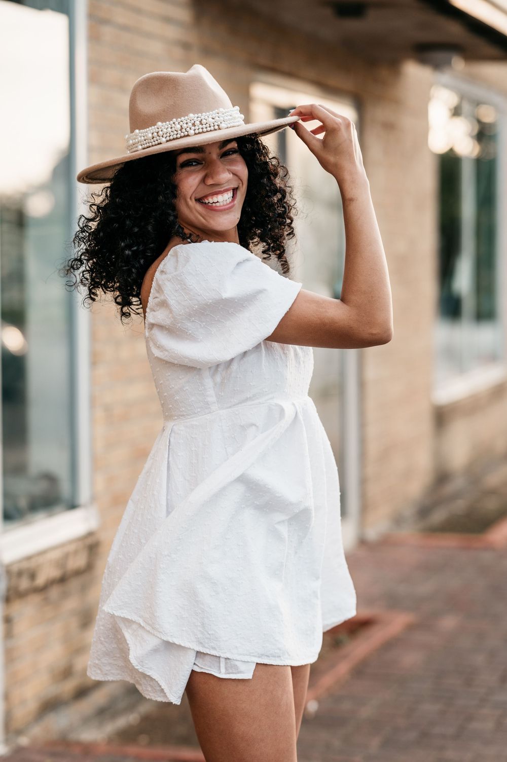 Young woman in white summer dress and brown hat poses playfully on a brick sidewalk in warm sunlight.