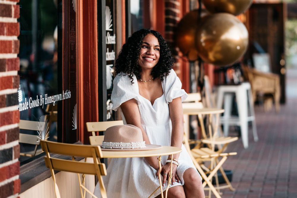 A person wearing a white dress smiles while sitting at an outdoor cafe with metal chairs and brick walls.