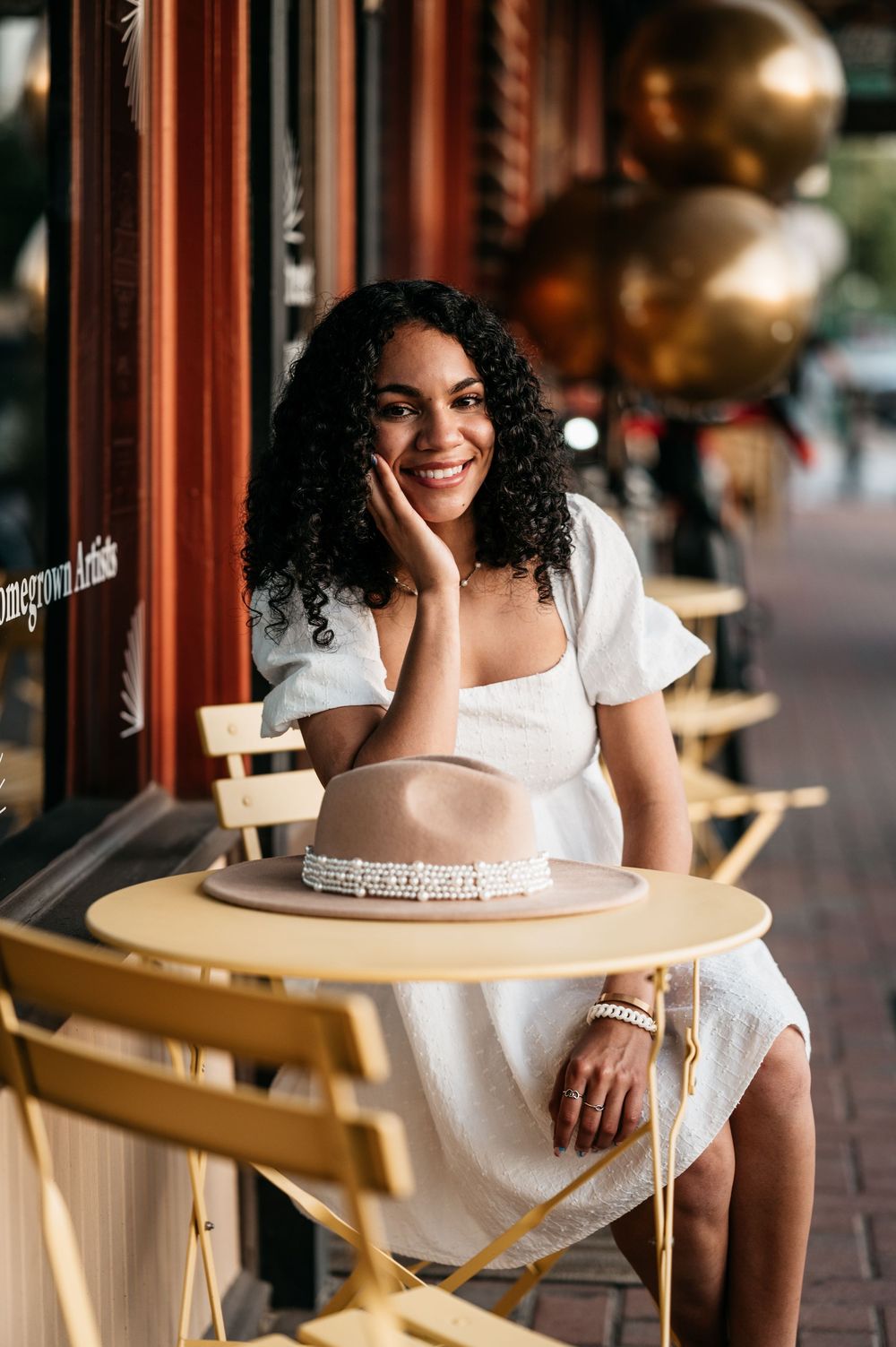 Smiling person with curly hair in white dress sitting at outdoor cafe table with copper decor in background.