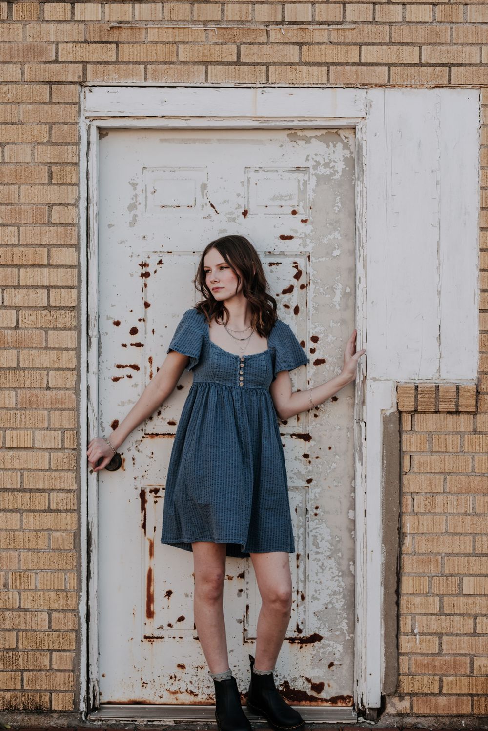 Woman in blue denim dress and boots poses against weathered white door and brick wall.