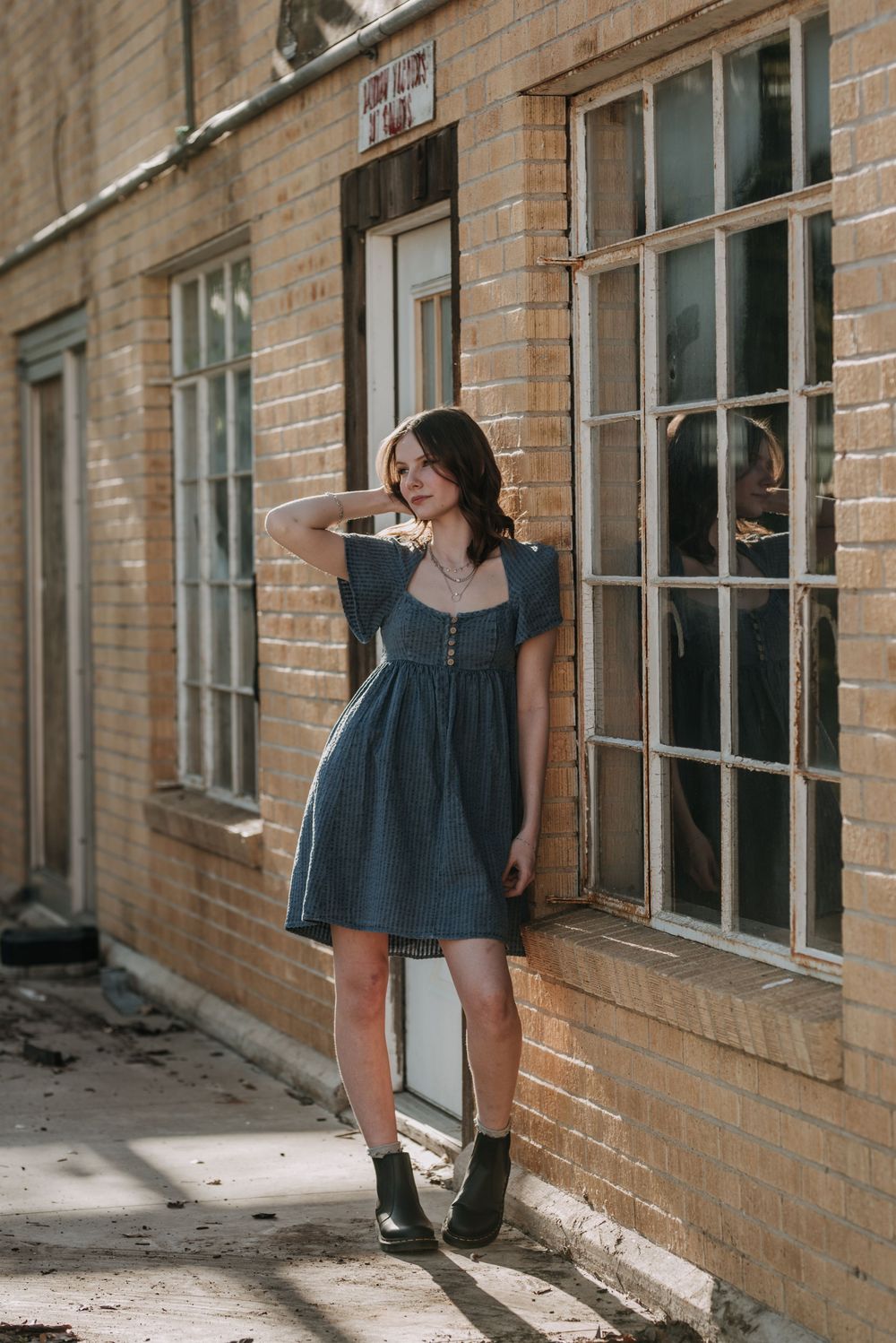 A person in a blue dress and boots poses against a brick wall near tall windows in natural lighting.