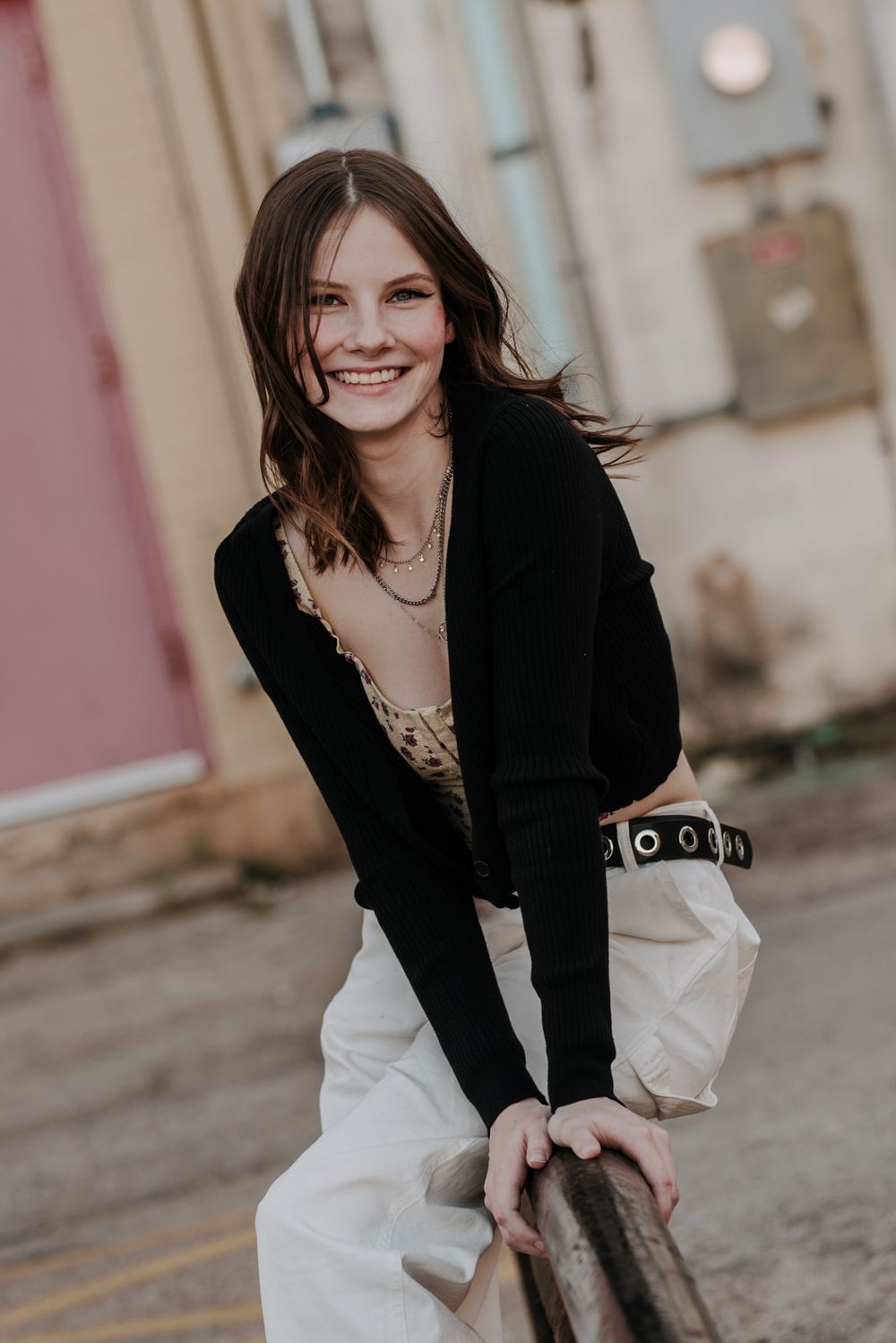 Casual portrait featuring a person in a black sweater and white pants leaning against a railing with a joyful expression.