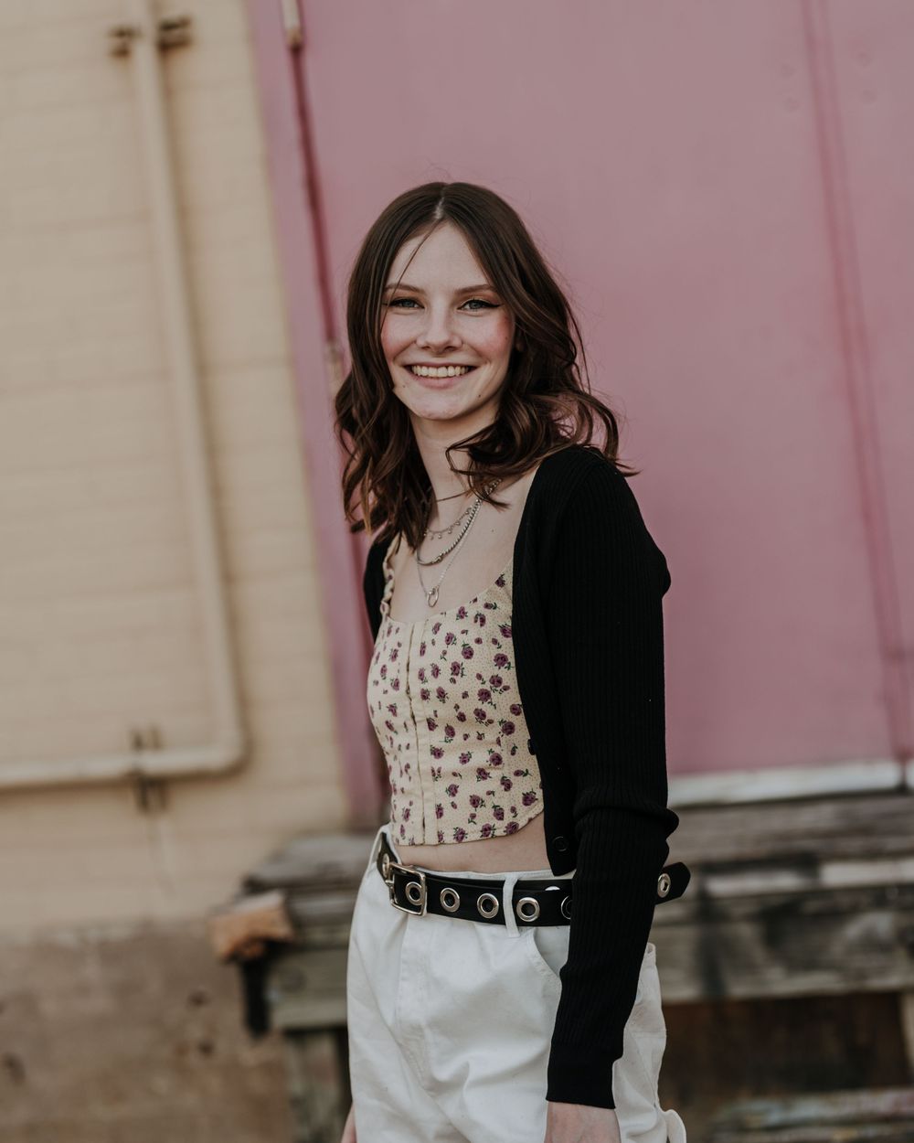 Young woman in a polka dot top and black cardigan smiles warmly against a pink and beige wall background.
