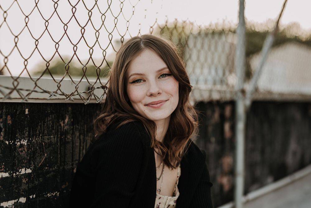 Portrait photo of someone with brown hair in front of a chain link fence, wearing a black top at sunset.