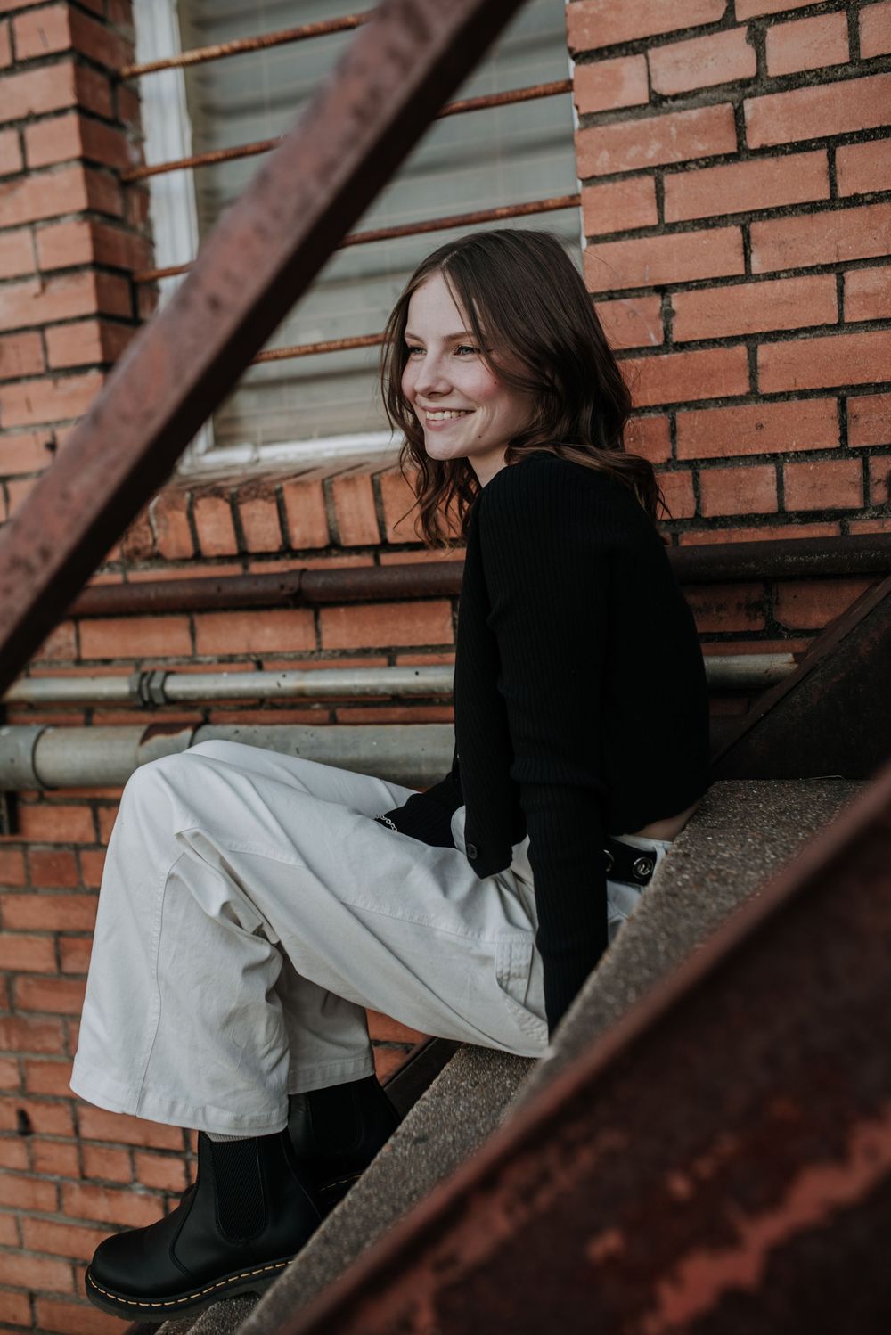 Portrait of someone in black sweater and beige pants sitting on brick steps with rustic industrial backdrop.