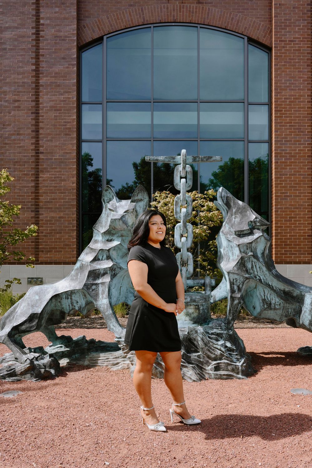 Graduate posing in front of decorative wolf statues and glass window wearing black dress and white shoes.