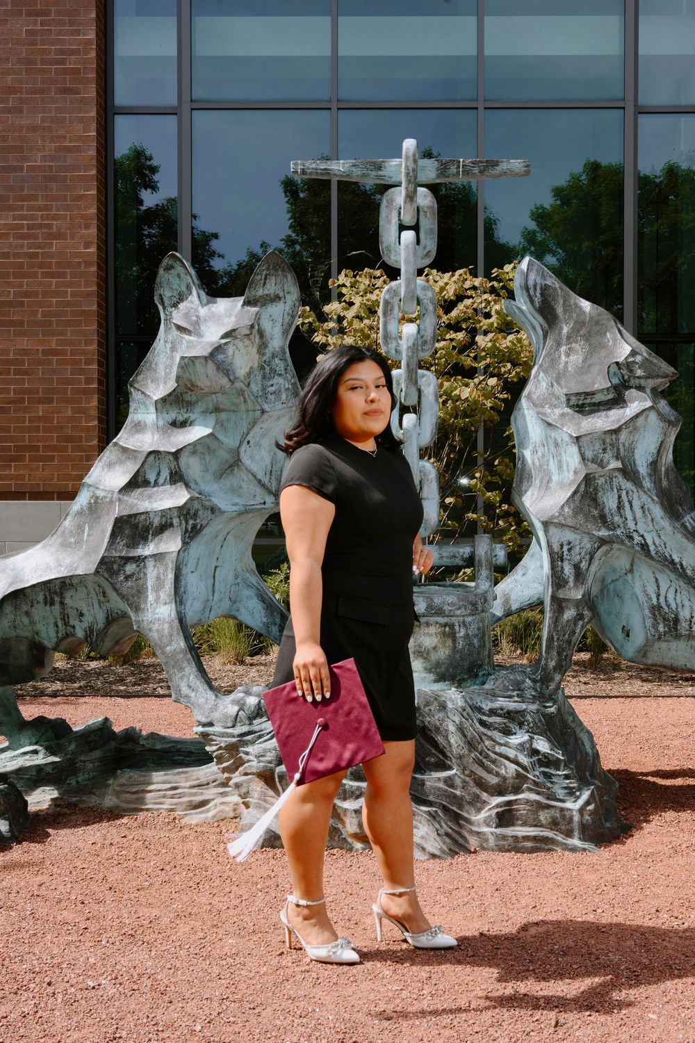 Graduate celebrates in a black dress and heels in front of a modern sculpture garden with reflective art pieces.