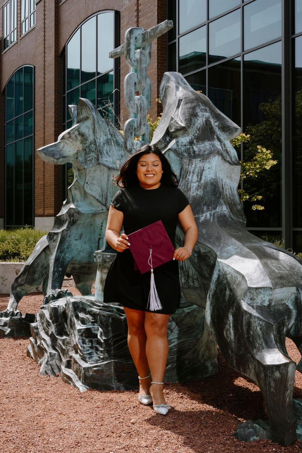 Graduate in black outfit holding diploma in front of modern sculptural art installation on campus.