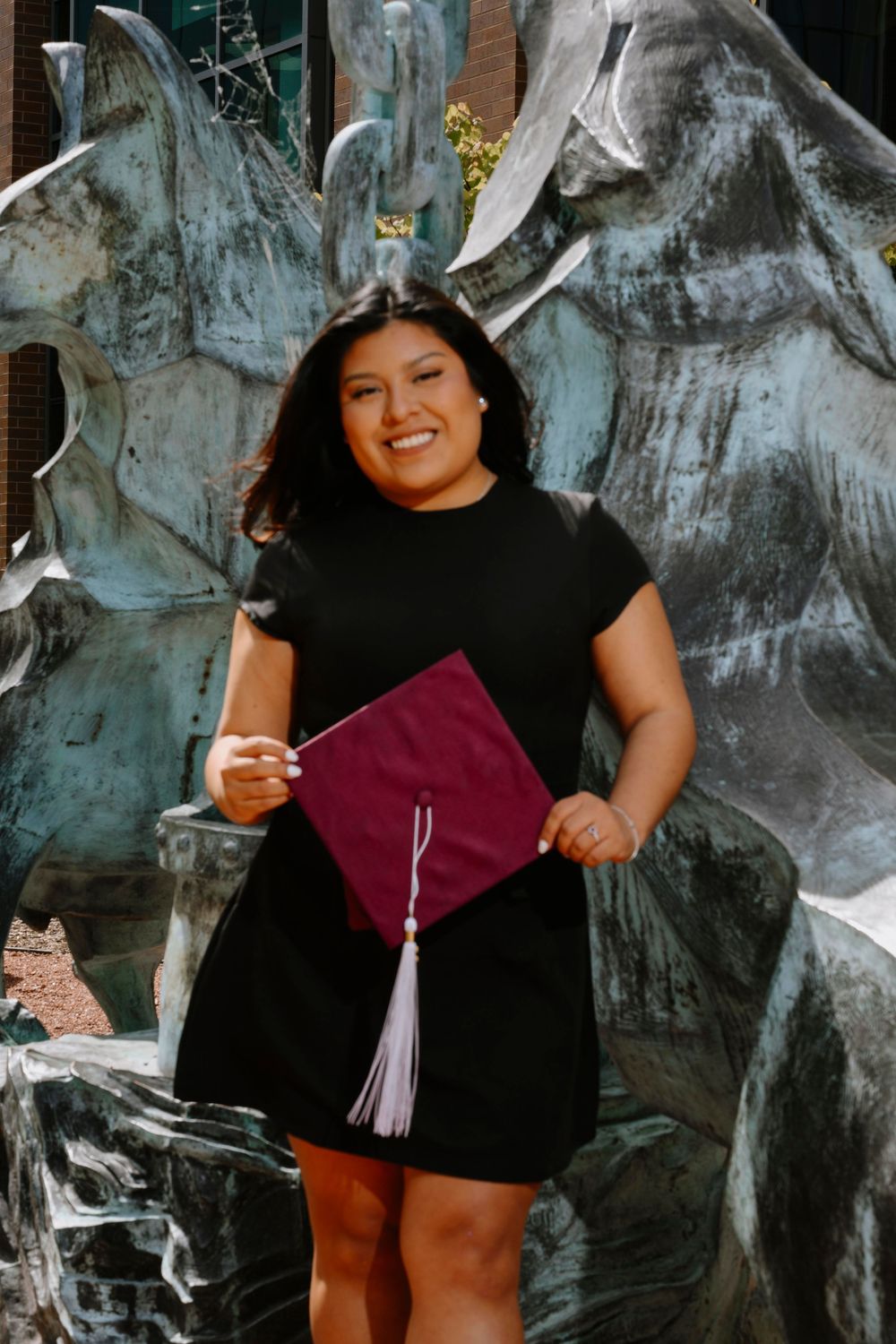 Graduate holding burgundy diploma cover while posing in front of metallic sculpture wearing black dress.