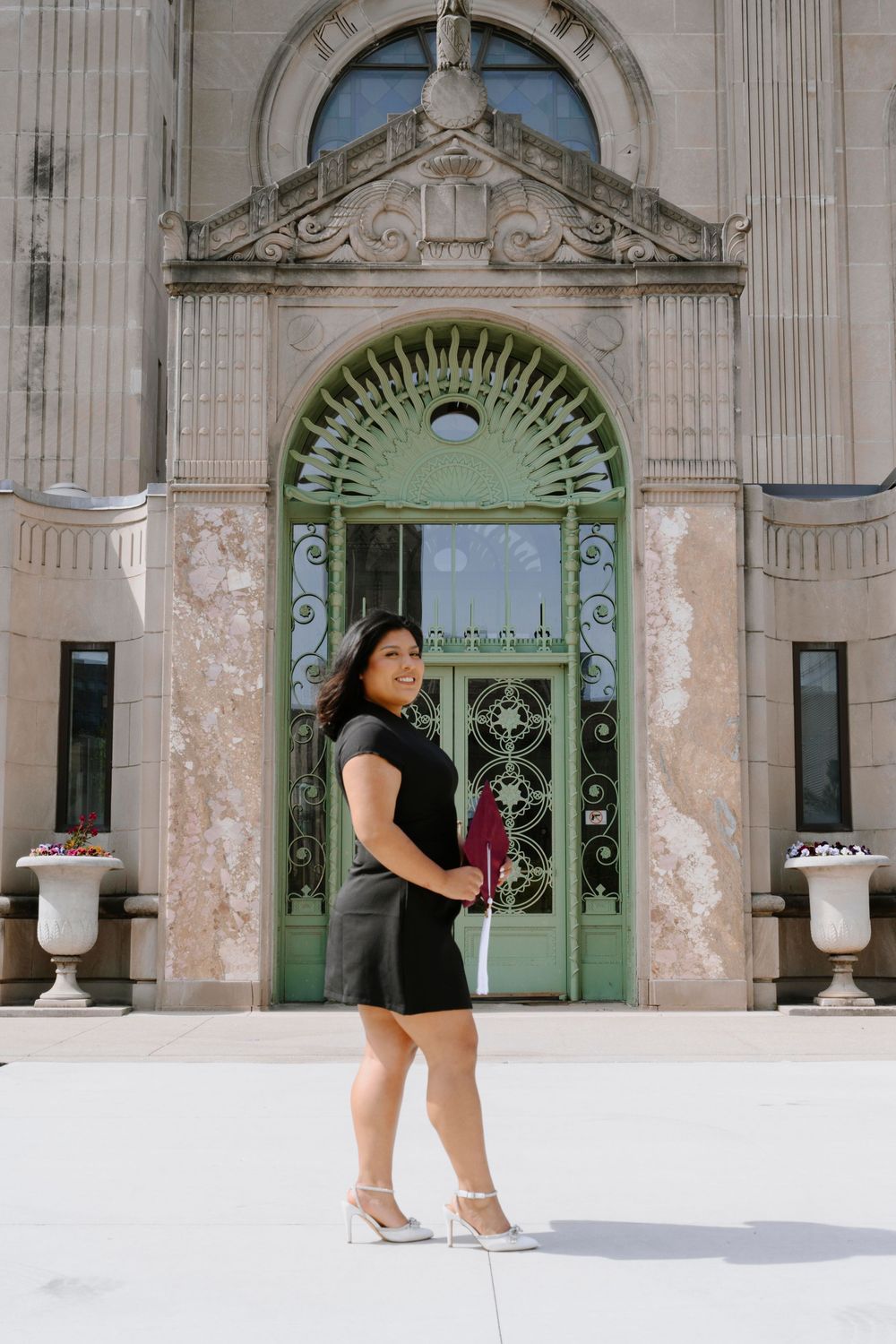 Graduate in a black dress poses in front of an ornate vintage doorway with decorative architectural details.