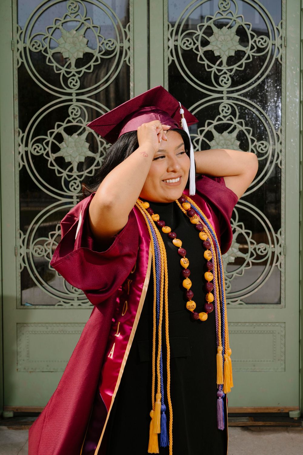 Graduate in burgundy cap and gown smiling and adjusting their tassel against ornate door background.