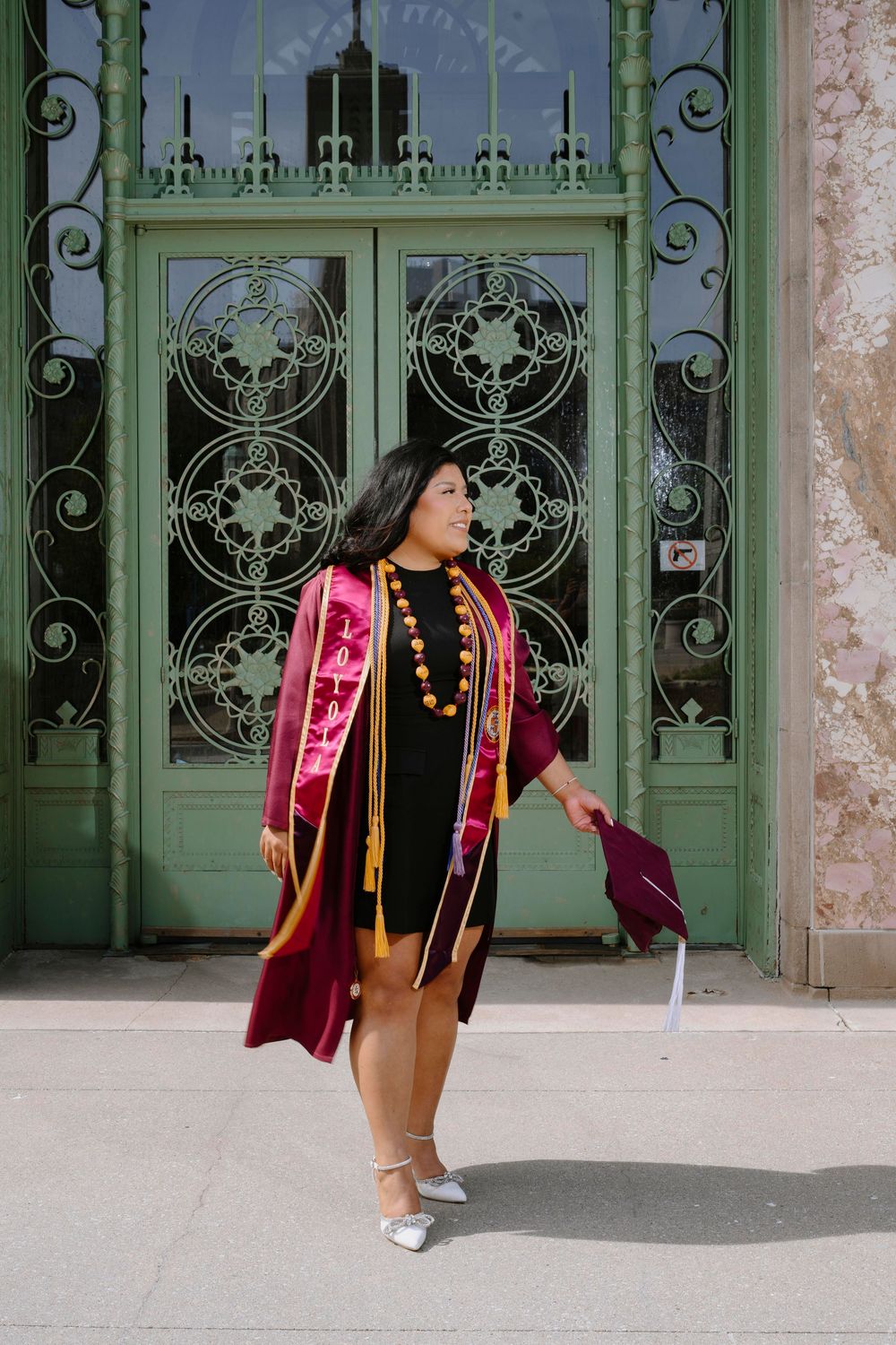 Graduate in academic regalia poses in front of ornate green doors wearing burgundy graduation gown.