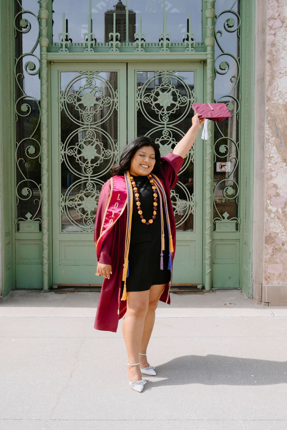 Graduate celebrates in burgundy graduation attire while posing joyfully in front of decorative green doors.