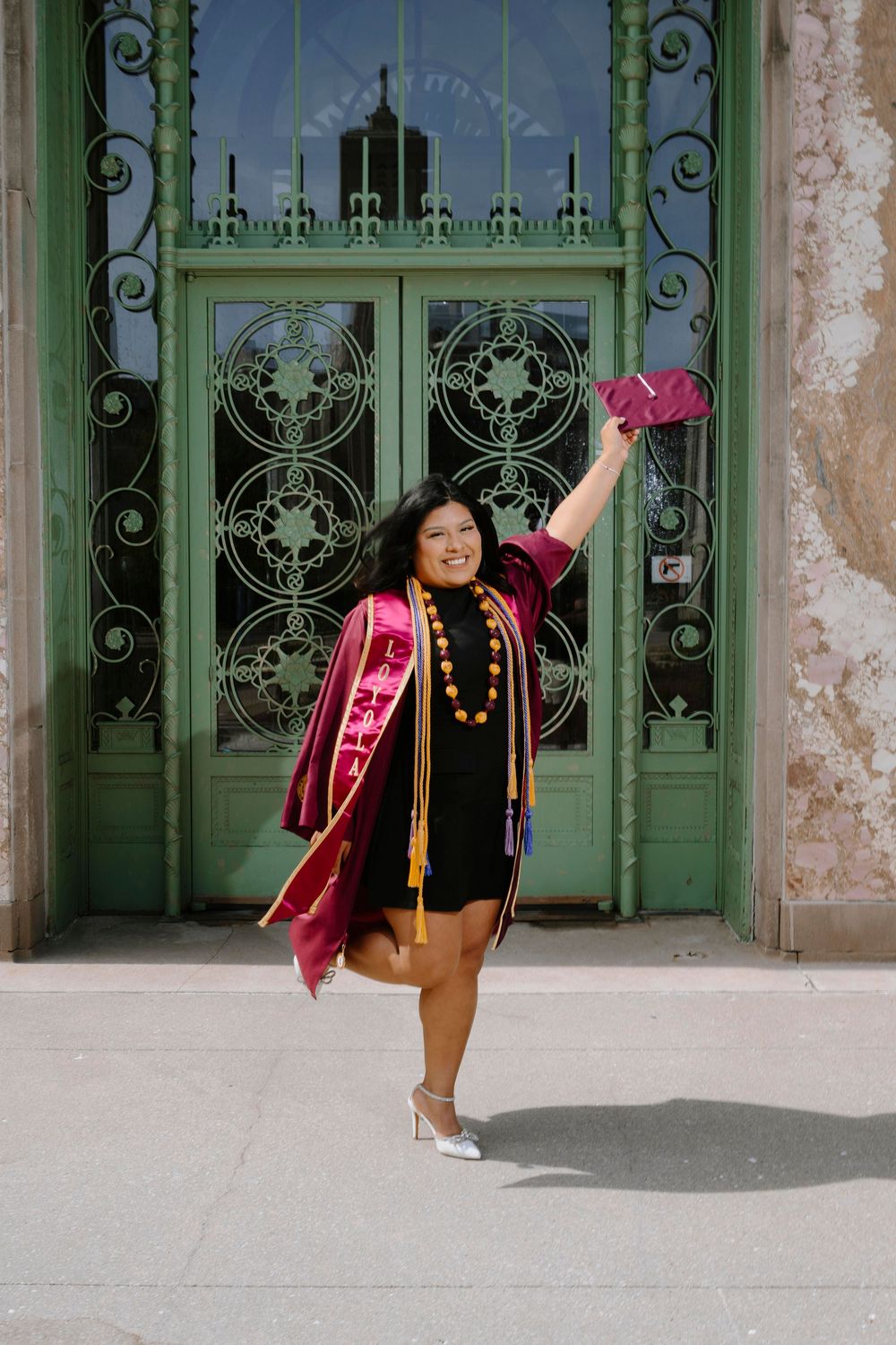 Graduate in academic regalia poses joyfully in front of decorative green doors wearing graduation stole and medallions.